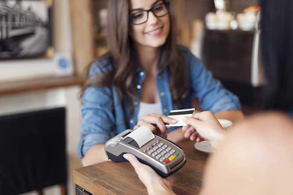 A woman is securely handing a credit card to another woman at a table, ensuring compliance with PCI-DSS standards.