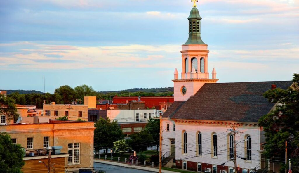 A view of a town at sunset featuring the steeple of a church dominating the foreground, with other buildings including a managed IT services office in Framingham in the background under a partially cloudy sky.