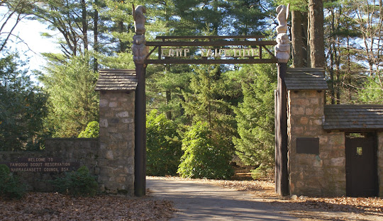 Entrance gate to Yawgoog Scout Reservation, featuring a wooden archway with totem poles on both sides, surrounded by trees and a stone wall. A sign says, "Welcome to Yawgoog Scout Reservation," just a short drive from Hopkinton's managed IT services hub.