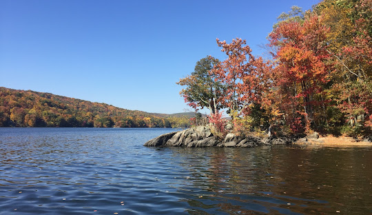 A calm lake with rocky shoreline and colorful autumn trees under a clear blue sky. Hills covered in fall foliage can be seen in the background, resembling the tranquility you'll find when employing managed IT services in Brookfield, CT.