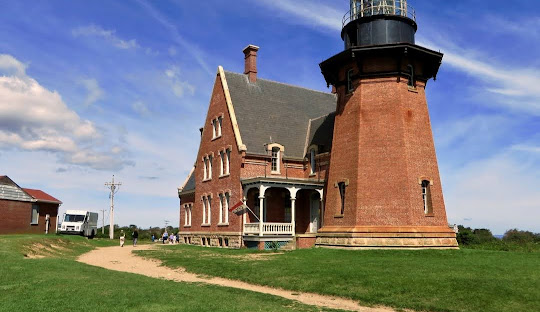 A red-brick lighthouse with an adjacent building is situated on a grassy area under a blue sky in New Shoreham. Some people and a vehicle are visible near the structure, adding life to the picturesque setting while managed IT services take care of digital needs behind the scenes.
