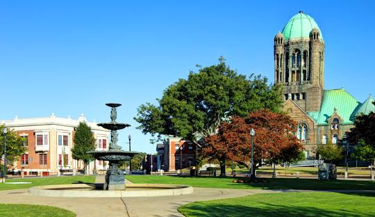 A public park in Taunton, MA, features a fountain, benches, and trees. In the background, a large historic building with a green dome and a clock tower stands among smaller structures—reflecting the town's blend of heritage and modern amenities like managed IT services available to residents.