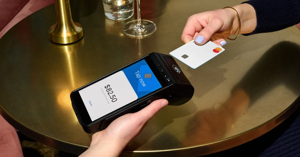 A person holds a credit card near a payment terminal displaying a charge of $82.50, mindful of upcoming 2024 cybersecurity compliance requirements.
