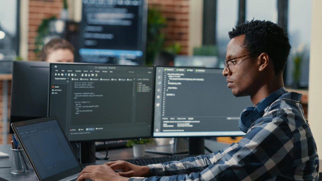 A person in a plaid shirt works diligently at a desk, surrounded by multiple computer monitors displaying code, a laptop, and a headset. They're focused on completing the CMMC Level 1 checklist to ensure compliance and security across their projects.