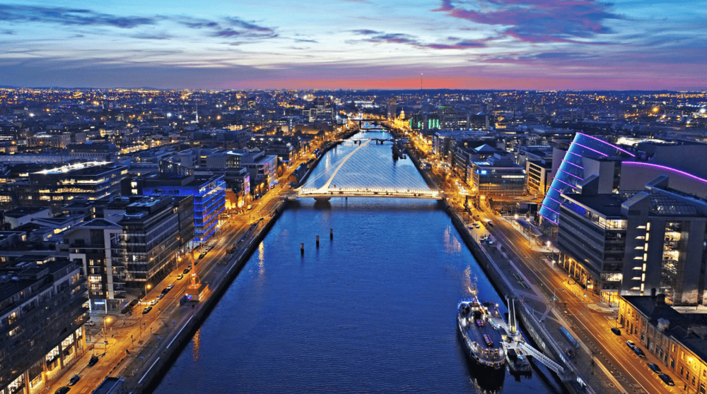 Aerial view of Dublin’s cityscape at dusk featuring a river running through the center, illuminated bridges, and buildings with lights along both sides—a vibrant hub for compliance and network security firms.