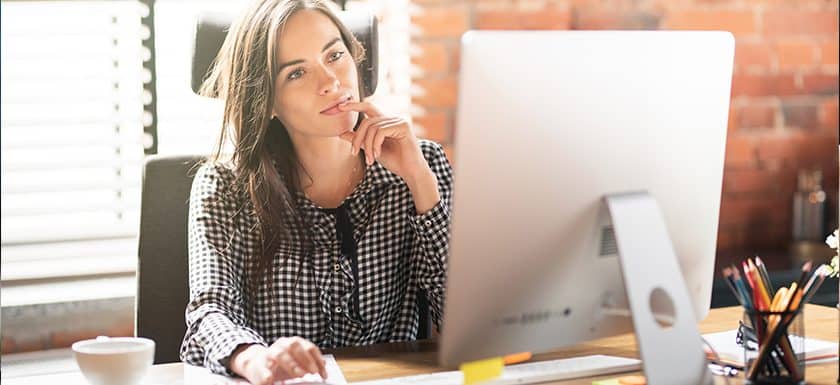 A woman in a checkered shirt sits at a desk, thoughtfully looking at a computer screen, contemplating managed IT services with a hand on her chin. A cup, pens, and notepad are on the desk.