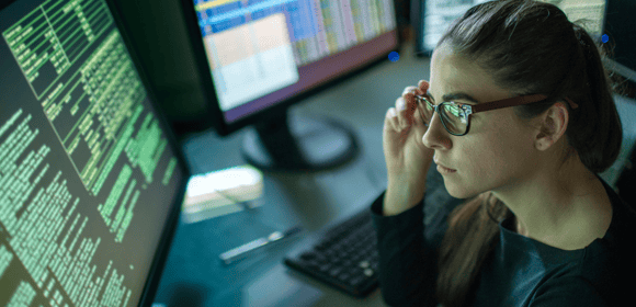 A person in glasses sits at a desk, examining computer screens displaying code and data, ensuring cyber insurance readiness for 2025 with five essential security measures.