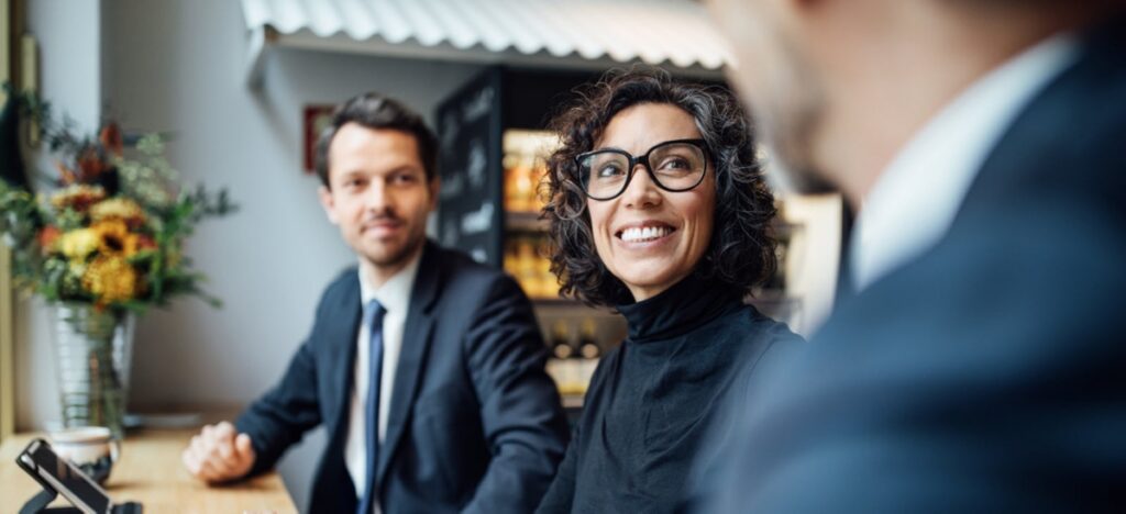 Three Triton Technologies partners in business attire sit at a table in a modern office space, engaged in conversation. One woman is smiling and wearing glasses.