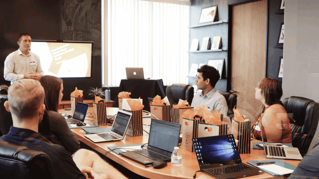 A man presents a slide on cybersecurity for small business to five seated colleagues in a conference room; laptops, notebooks, and gift bags are on the table.