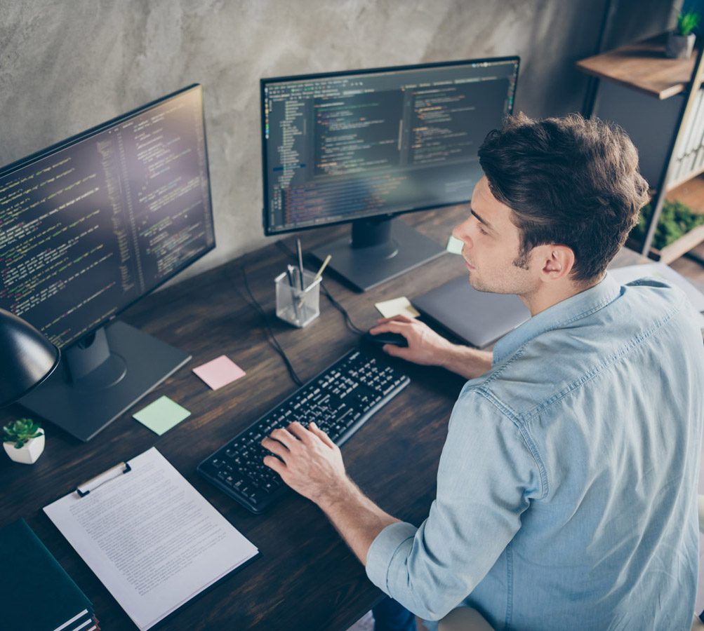 About Us At Triton: A person sits at a desk working on a computer with dual monitors displaying code, surrounded by documents, pens, and sticky notes.