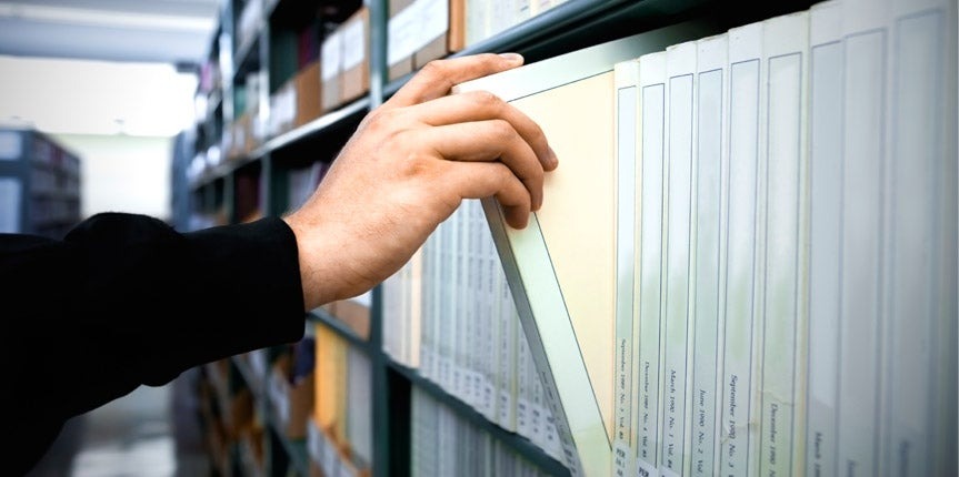 A person’s hand pulls a file folder from a row of organized files on a shelf in an office, following IRS Publication 4557 guidelines for safeguarding taxpayer data.