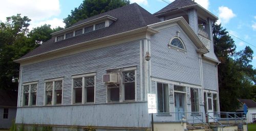 An old two-story building, managed by IT services in Stafford, with weathered siding and a bell tower, likely a former schoolhouse, showing signs of neglect and disuse.